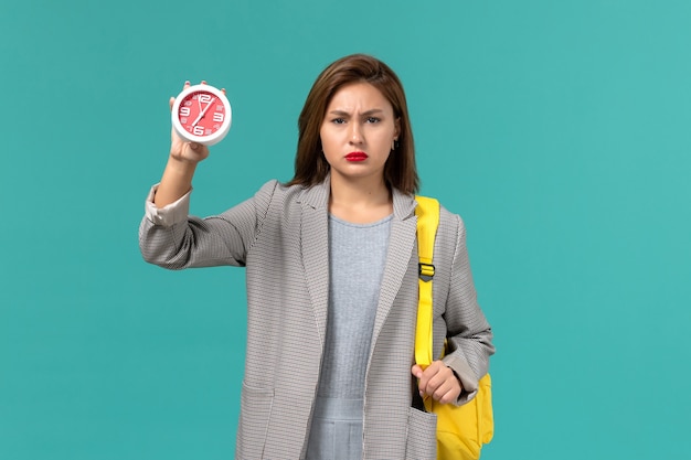 Front view of female student in grey jacket wearing her yellow backpack holding clocks on blue wall