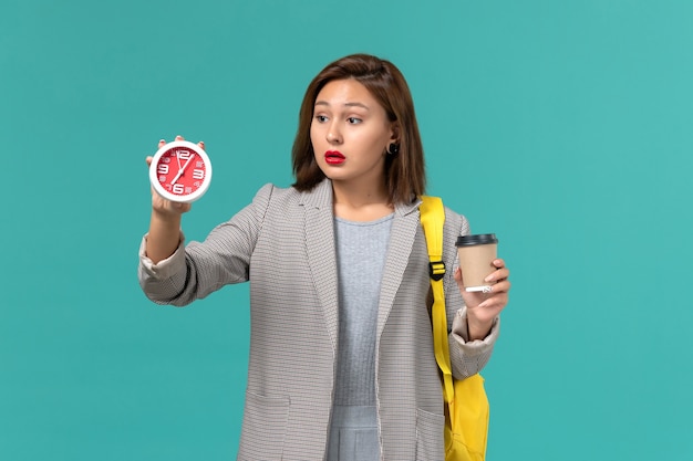 Free photo front view of female student in grey jacket wearing her yellow backpack holding clocks and coffee on blue wall