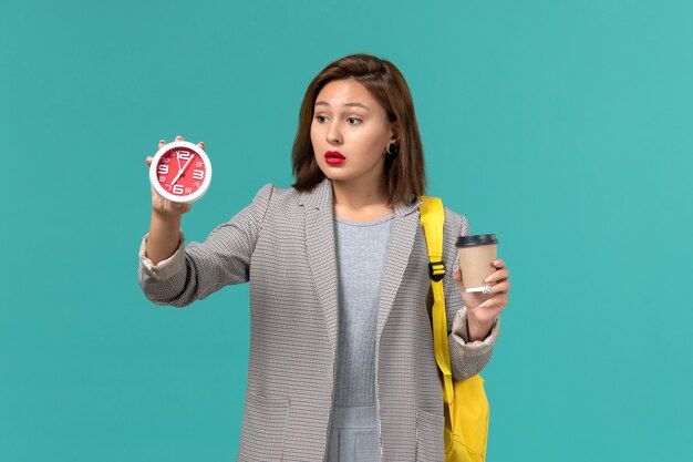 Front view of female student in grey jacket wearing her yellow backpack holding clocks and coffee on blue wall