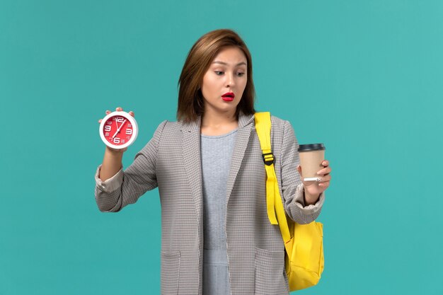 Front view of female student in grey jacket wearing her yellow backpack holding clocks and coffee on blue wall