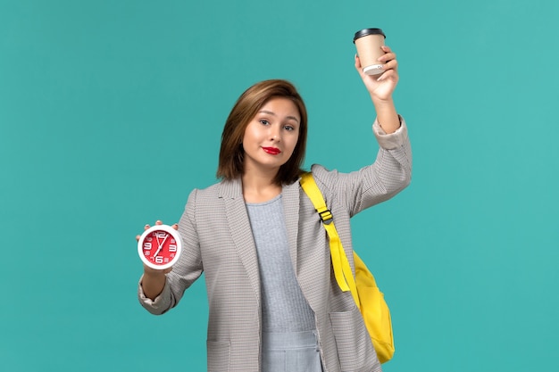 Front view of female student in grey jacket wearing her yellow backpack holding clocks and coffee on blue wall