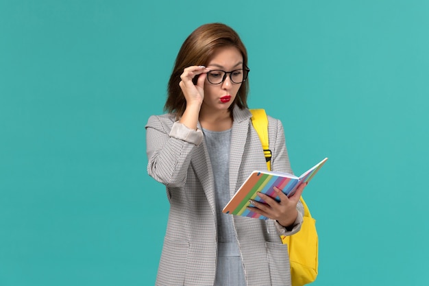 Front view of female student in grey jacket wearing her yellow backpack holding copybook on light blue wall