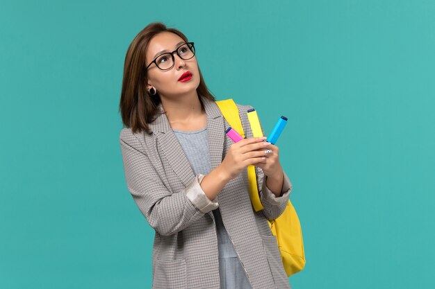 Front view of female student in grey jacket wearing her yellow backpack holding felt pens on light blue wall