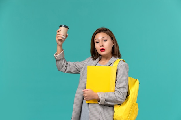 水色の壁にファイルとコーヒーを保持している彼女の黄色のバックパックを身に着けている灰色のジャケットの女子学生の正面図