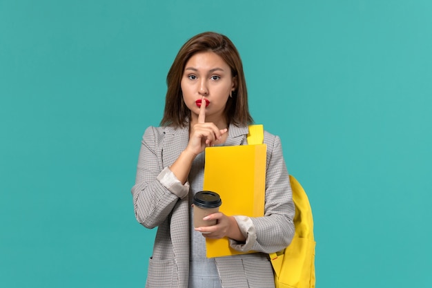 Front view of female student in grey jacket wearing her yellow backpack holding files and coffee on light-blue wall