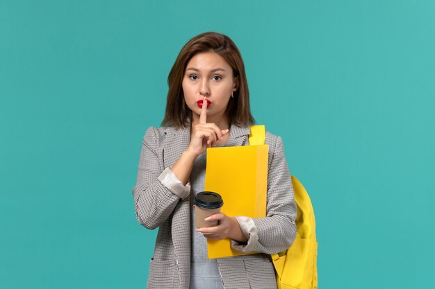 Front view of female student in grey jacket wearing her yellow backpack holding files and coffee on light-blue wall