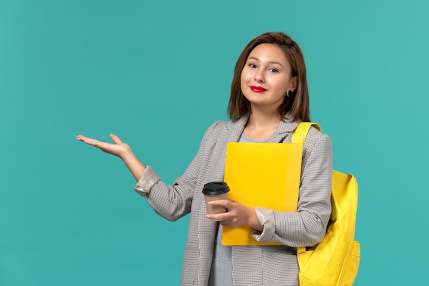 Front view of female student in grey jacket wearing her yellow backpack holding files and coffee on light-blue wall