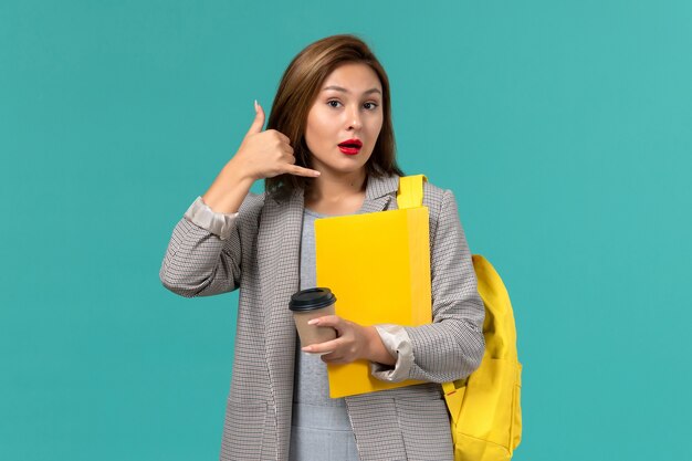 Front view of female student in grey jacket wearing her yellow backpack holding files and coffee on light blue wall