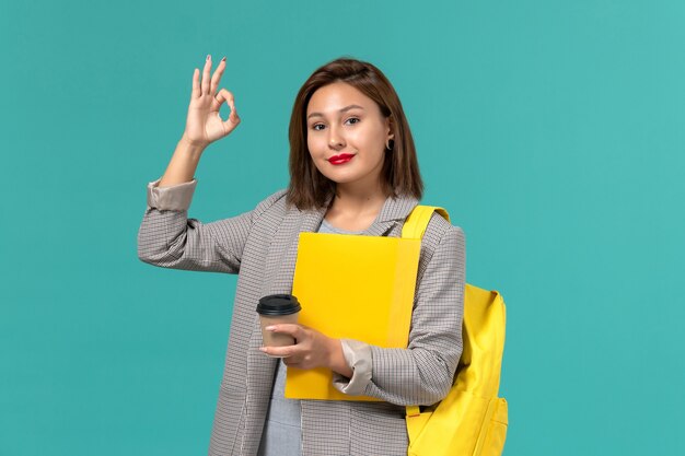 Front view of female student in grey jacket wearing her yellow backpack holding files and coffee smiling on light-blue wall