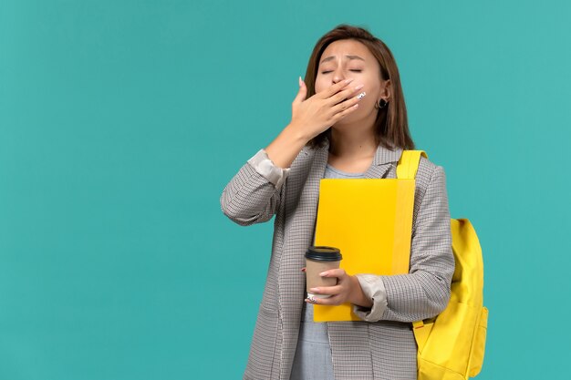Front view of female student in grey jacket wearing her yellow backpack holding files and coffee yawning on light-blue wall