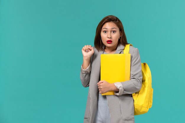 그녀의 노란색 배낭을 착용하고 밝은 파란색 벽에 파일을 들고 회색 재킷을 입은 여성 학생의 전면보기