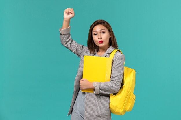 Front view of female student in grey jacket wearing her yellow backpack and holding files on light-blue wall