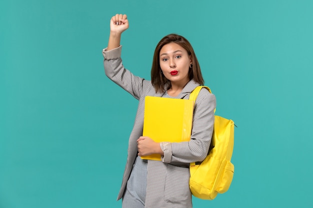 Front view of female student in grey jacket wearing her yellow backpack and holding files on light-blue wall