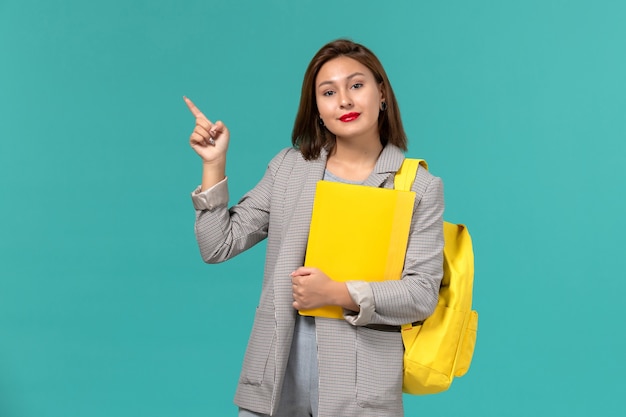 Front view of female student in grey jacket wearing her yellow backpack and holding files on light-blue wall