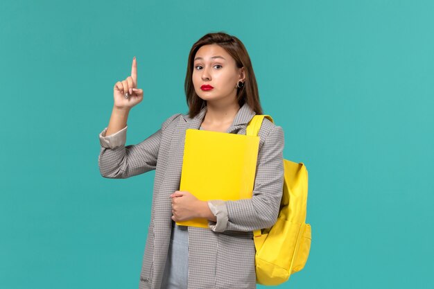 Front view of female student in grey jacket wearing her yellow backpack and holding files on light-blue wall