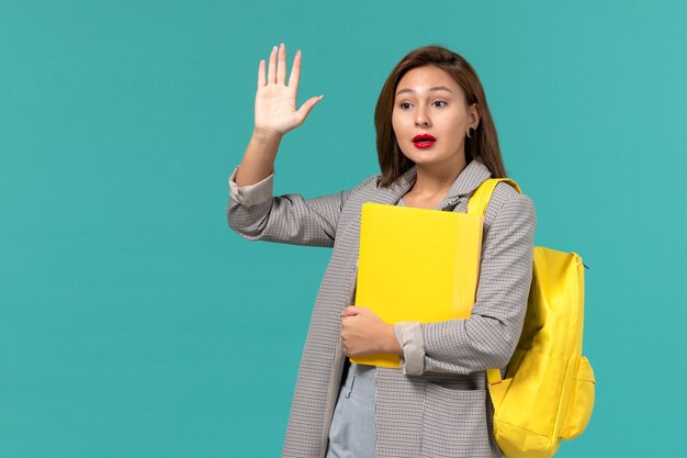 Front view of female student in grey jacket wearing her yellow backpack and holding files on light blue wall