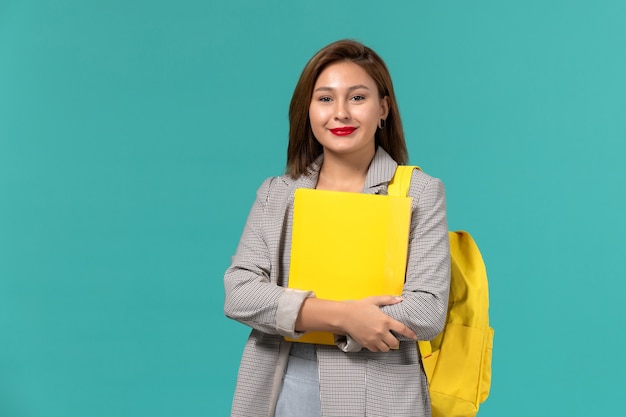Front view of female student in grey jacket wearing her yellow backpack and holding files on light blue wall