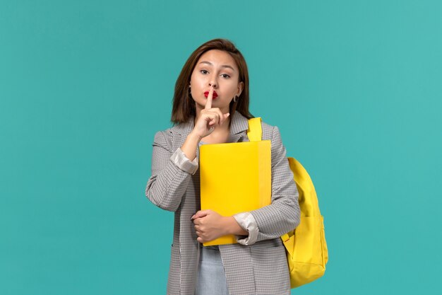 Front view of female student in grey jacket wearing her yellow backpack and holding files on the light blue wall