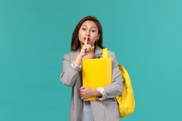 Front view of female student in grey jacket wearing her yellow backpack and holding files on the light blue wall