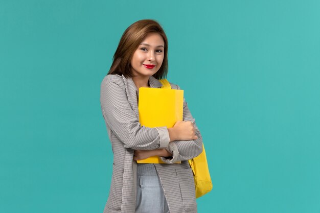 Front view of female student in grey jacket wearing her yellow backpack and holding files on the light blue wall