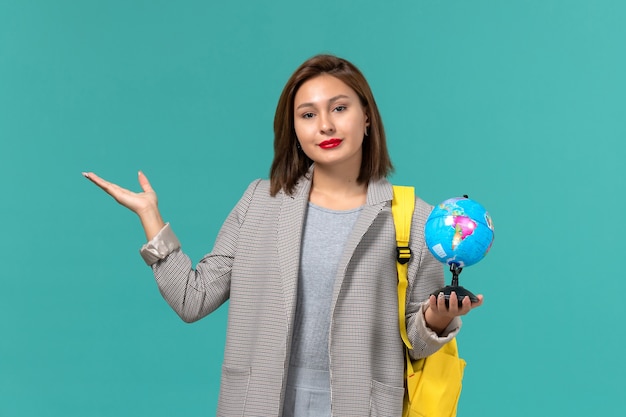 Front view of female student in grey jacket wearing her yellow backpack holding little globe on light blue wall