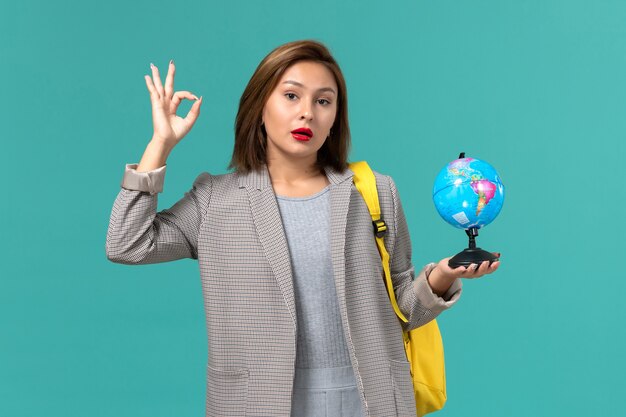 Front view of female student in grey jacket wearing her yellow backpack holding little globe on light blue wall