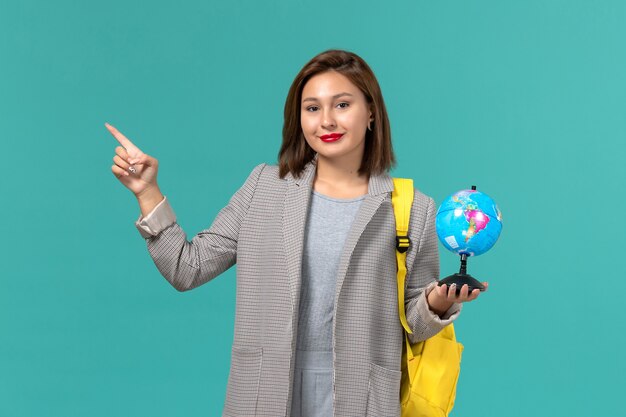 Front view of female student in grey jacket wearing her yellow backpack holding little globe on light blue wall