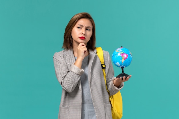 Front view of female student in grey jacket wearing her yellow backpack holding little globe thinking on light blue wall