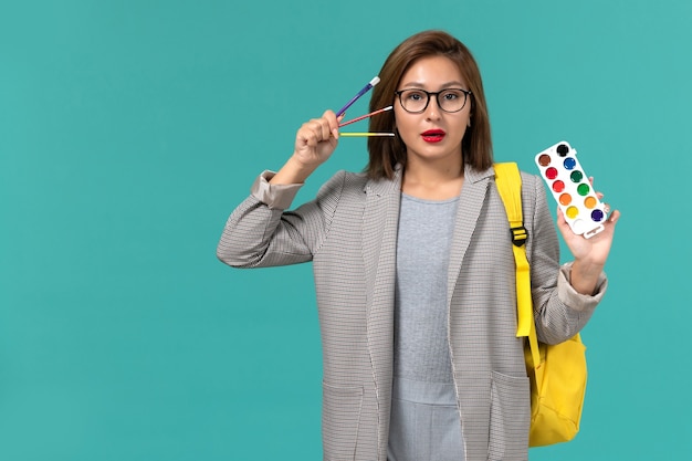 Front view of female student in grey jacket wearing her yellow backpack holding paints on blue wall