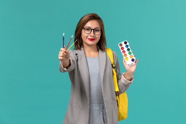 Front view of female student in grey jacket wearing her yellow backpack holding paints on the blue wall