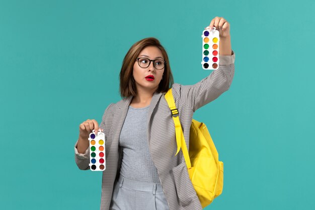 Front view of female student in grey jacket wearing her yellow backpack holding paints for drawing on blue wall