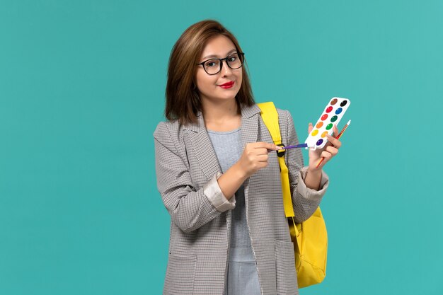 Front view of female student in grey jacket wearing her yellow backpack holding paints smiling on blue wall