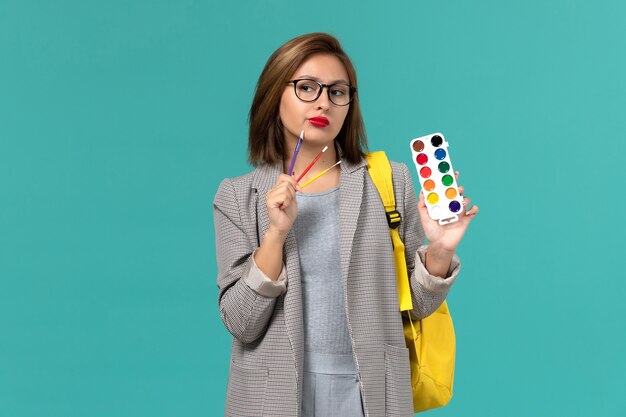 Front view of female student in grey jacket wearing her yellow backpack holding paints thinking on blue wall