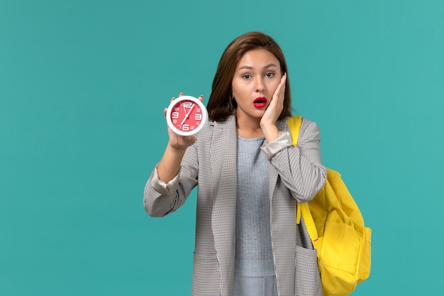 Front view of female student in grey jacket wearing yellow backpack holding clocks on the light blue wall