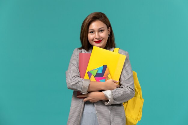 Front view of female student in grey jacket wearing yellow backpack holding copybook and files on the light blue wall