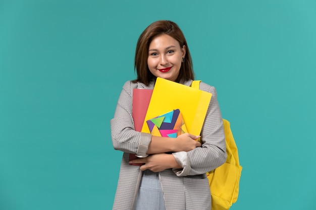 Front view of female student in grey jacket wearing yellow backpack holding copybook and files on the light blue wall