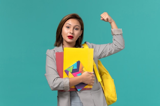 Front view of female student in grey jacket wearing yellow backpack holding copybook and files on the light-blue wall