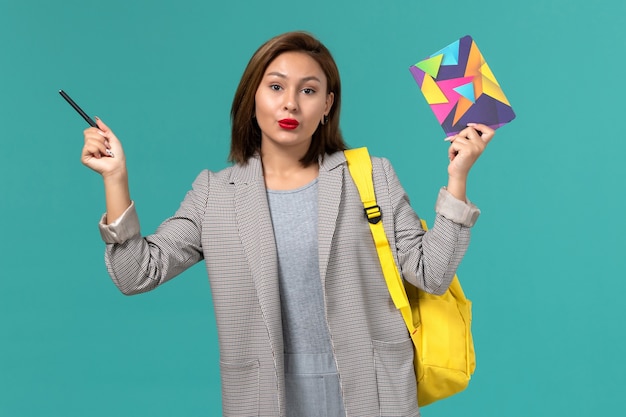 Front view of female student in grey jacket wearing yellow backpack holding copybook on the light-blue wall