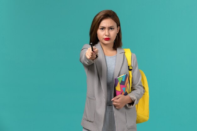 Front view of female student in grey jacket wearing yellow backpack holding copybook with pen on blue surface