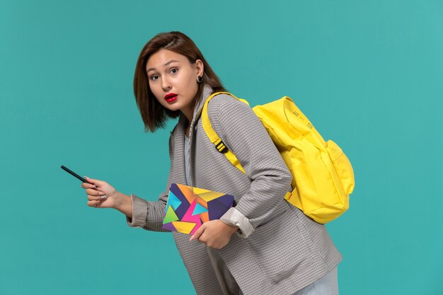 Front view of female student in grey jacket wearing yellow backpack holding copybook with pen on light-blue wall