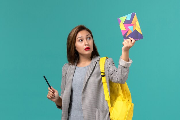 Front view of female student in grey jacket wearing yellow backpack holding copybook with pen on the light-blue wall