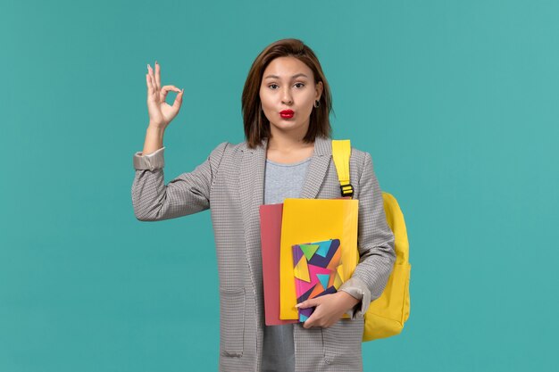 Front view of female student in grey jacket wearing yellow backpack holding files and copybook on blue wall