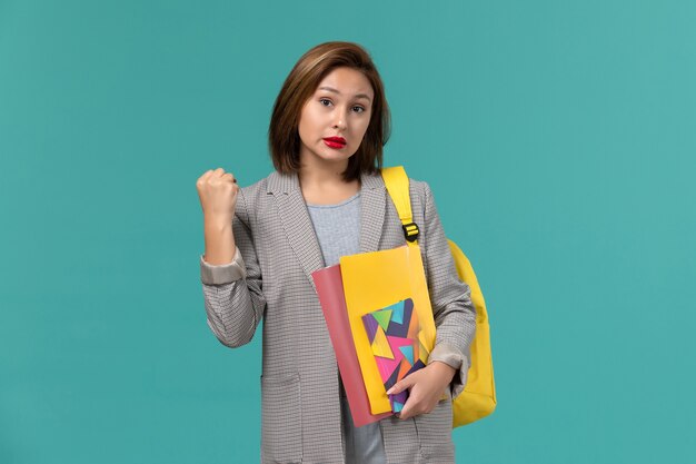 Front view of female student in grey jacket wearing yellow backpack holding files and copybook on the blue wall