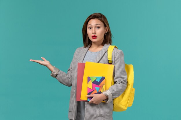 Front view of female student in grey jacket wearing yellow backpack holding files and copybook on the blue wall