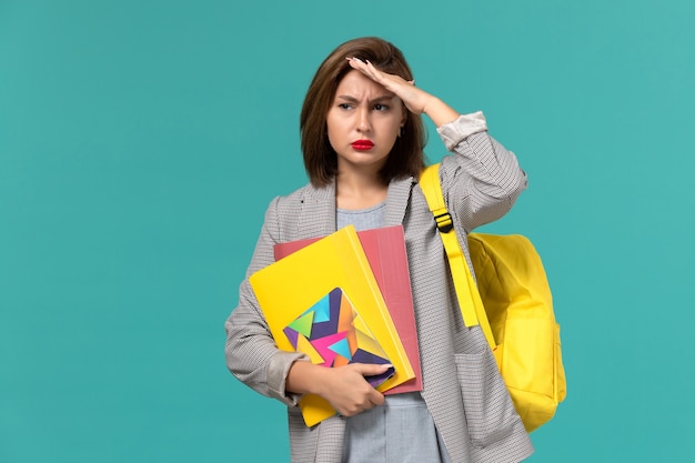 Front view of female student in grey jacket wearing yellow backpack holding files and copybook confused on blue wall