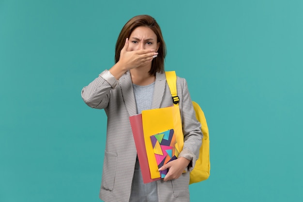 Front view of female student in grey jacket wearing yellow backpack holding files and copybook covering her mouth on blue wall