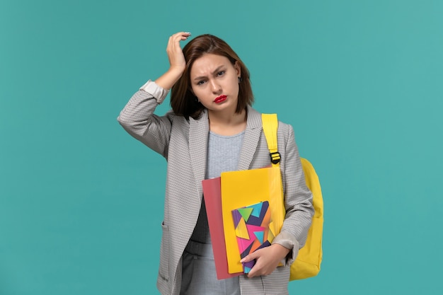 ファイルと青い壁に頭痛を持っているコピーブックを保持している黄色のバックパックを身に着けている灰色のジャケットの女子学生の正面図