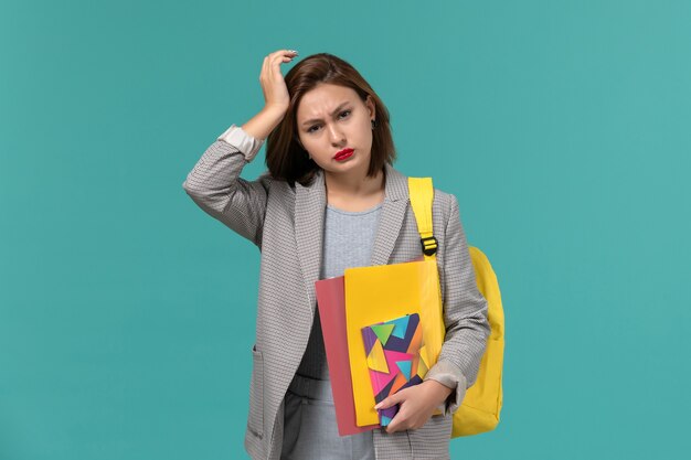 Front view of female student in grey jacket wearing yellow backpack holding files and copybook having headache on blue wall