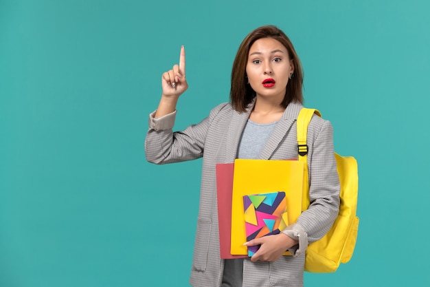 Front view of female student in grey jacket wearing yellow backpack holding files and copybook on light-blue wall