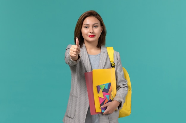 Front view of female student in grey jacket wearing yellow backpack holding files and copybook smiling on blue wall